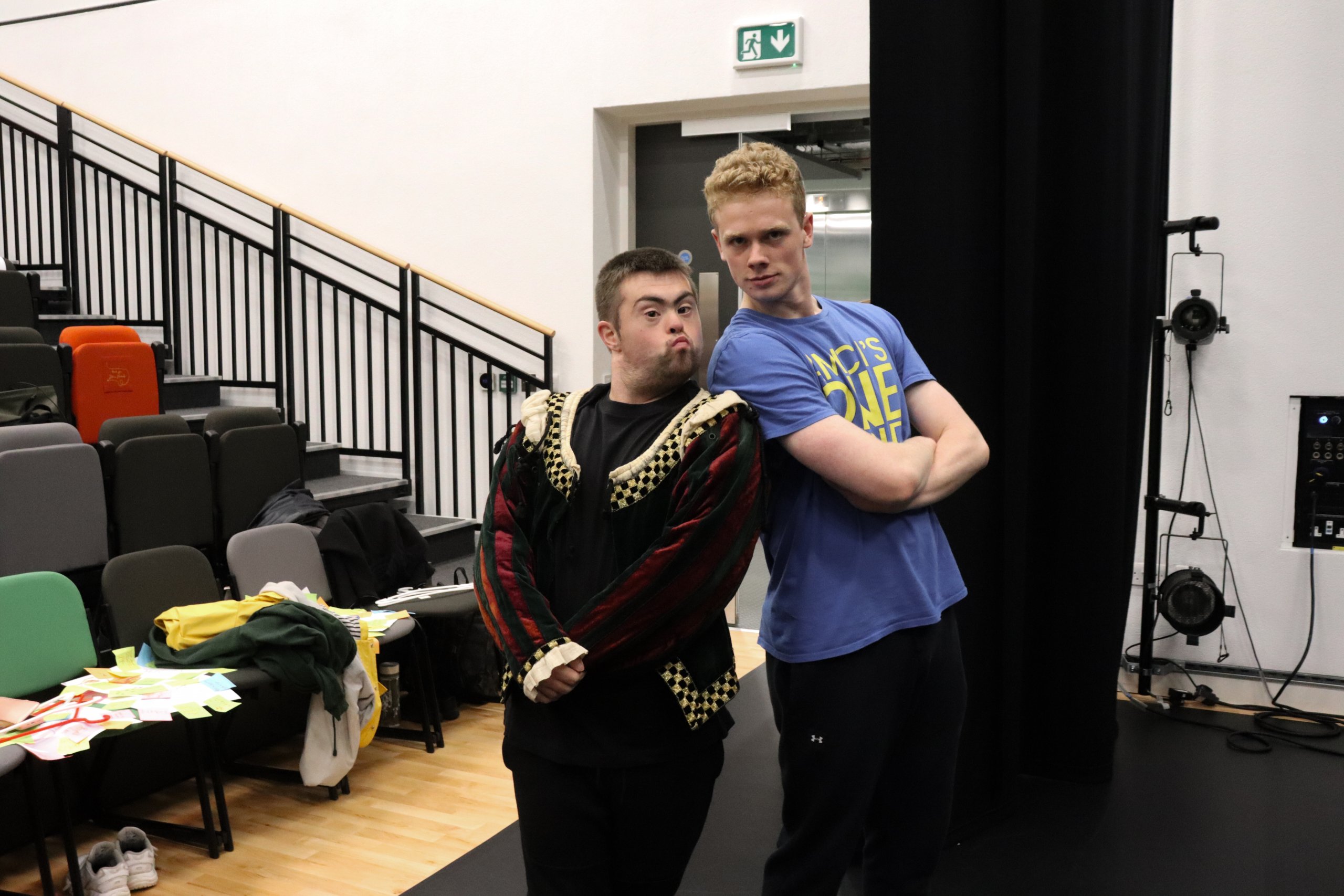 Two dancers standing back to back with arms folded in front of them. One dancer is in a costume. 