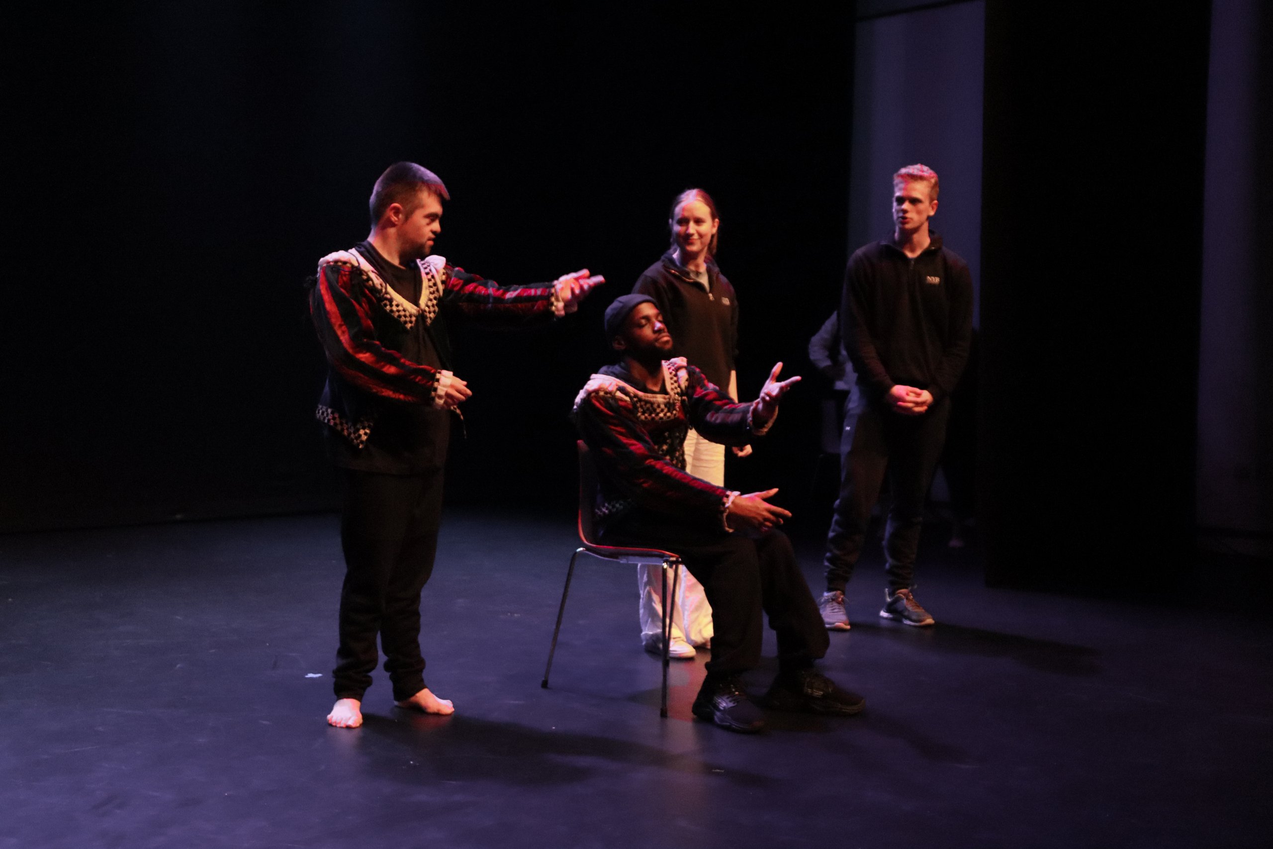 Two dancers in costume, one standing and one seated, both have their arms extended out. There are two people behind them observing this. 