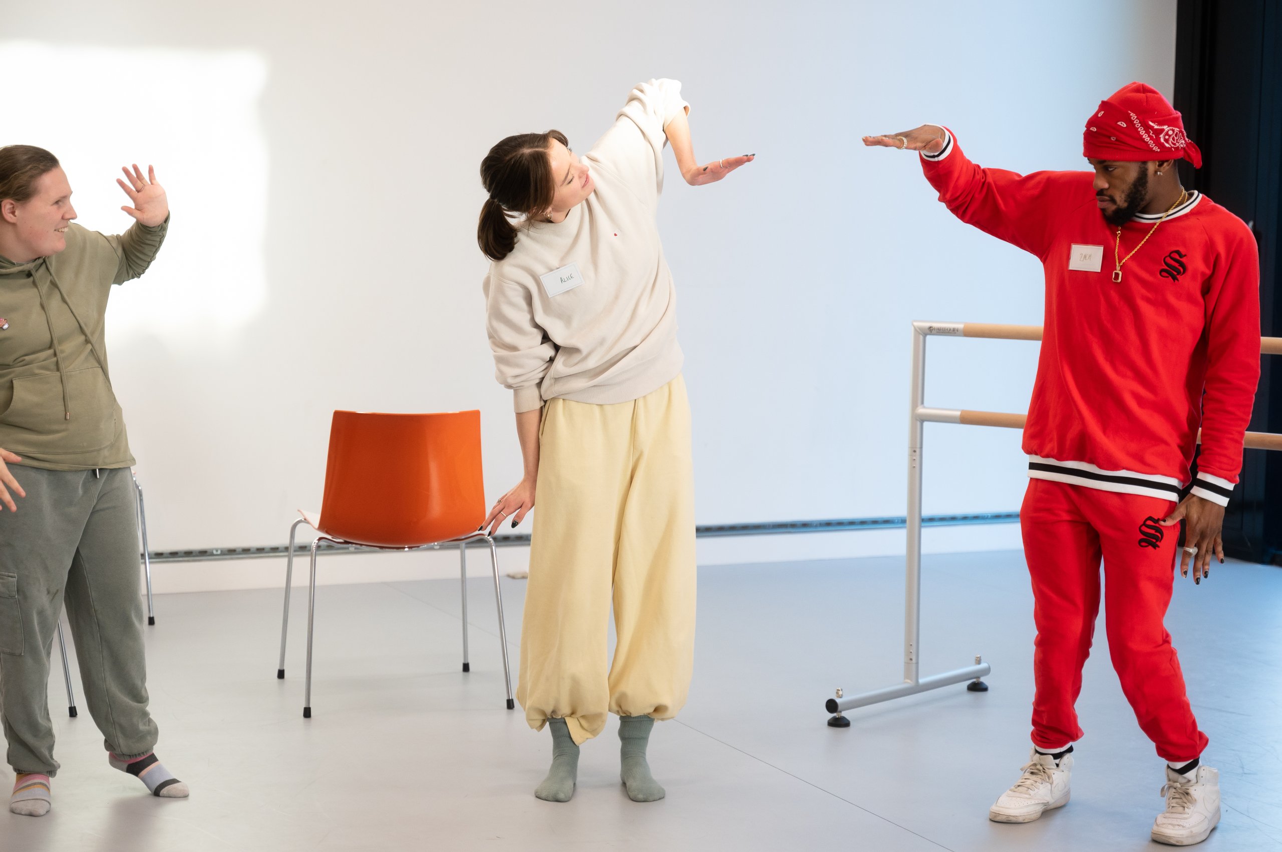 Three dancers standing. Two dancers are lifting an bent arm with a flexed wrist and the other dancer is lifting one arm showing their palm with fingers spread. There is a ballet barre and a red chair behind the dancers. 