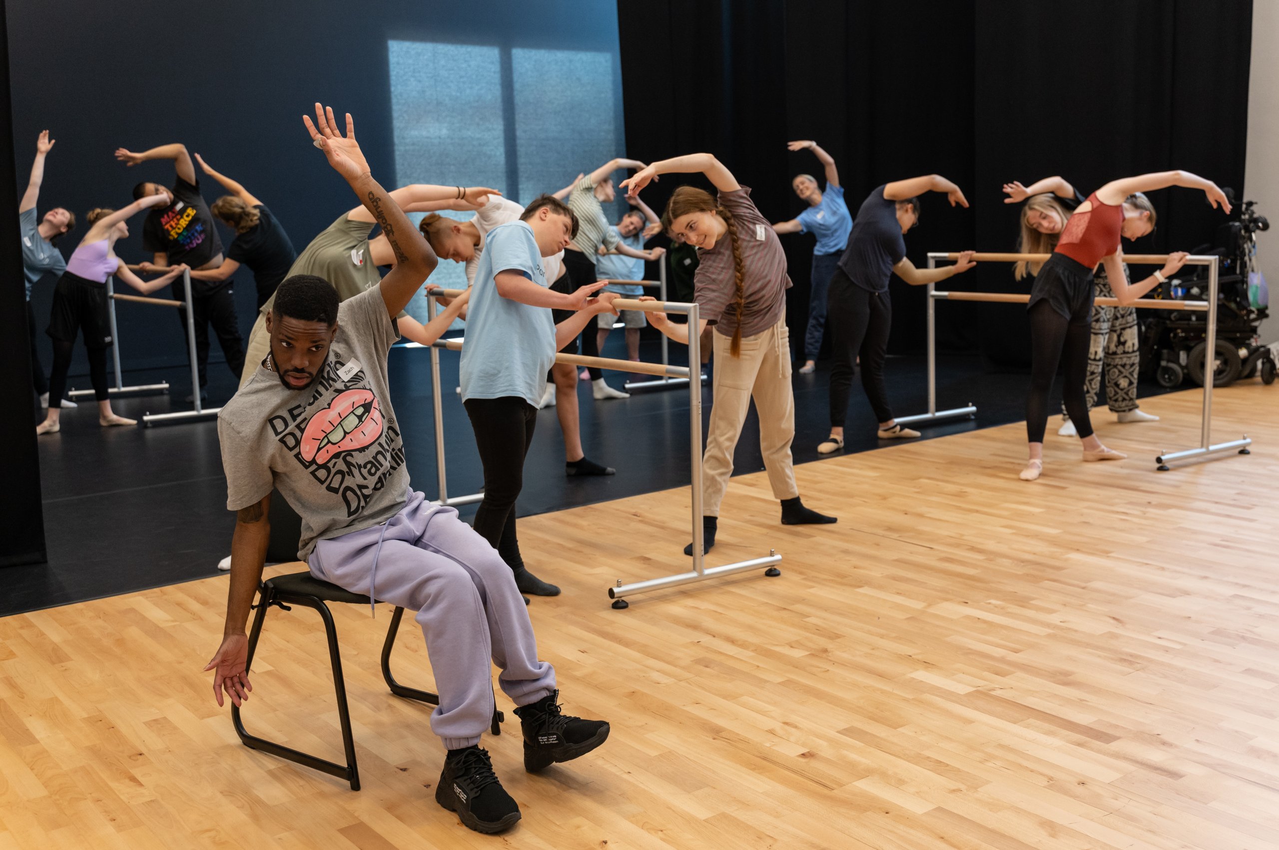 Many dancers holding onto a ballet barre with feet in second position, curving over towards the barre. One dancer is seated on a chair also reaching overhead. 
