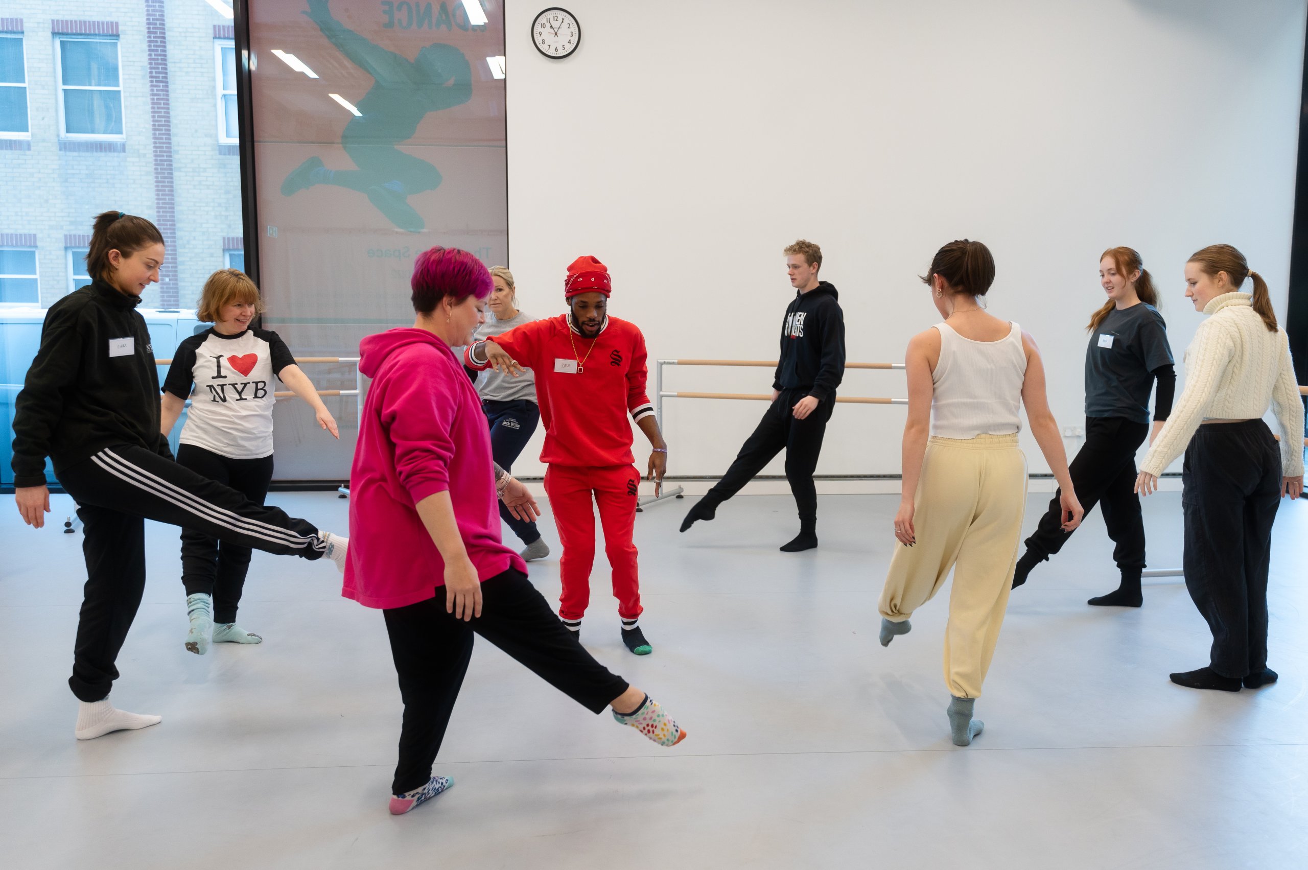 Nine dancers facing different directions with arms placed by their side, either lifting one leg off the floor or with two feet on the ground. 