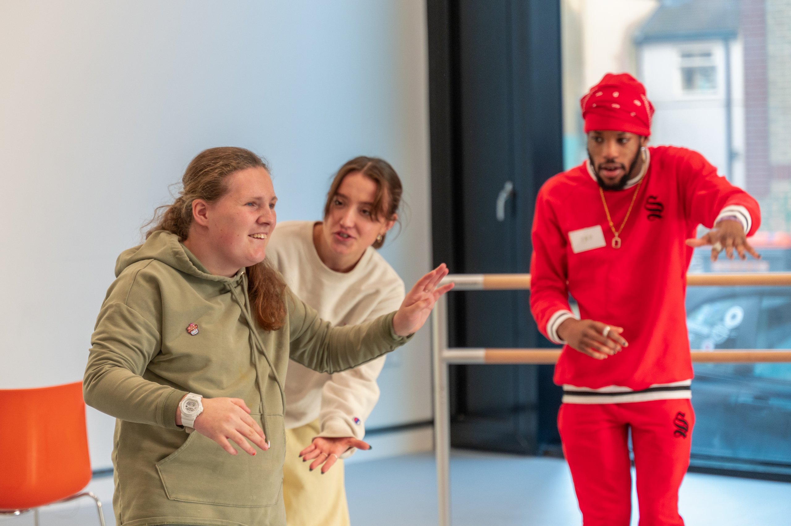 A dancer dressed in all red with one arm slightly lifted in front of them. Two dancers are interacting with one place slightly behind the other, both with arms in motion. 