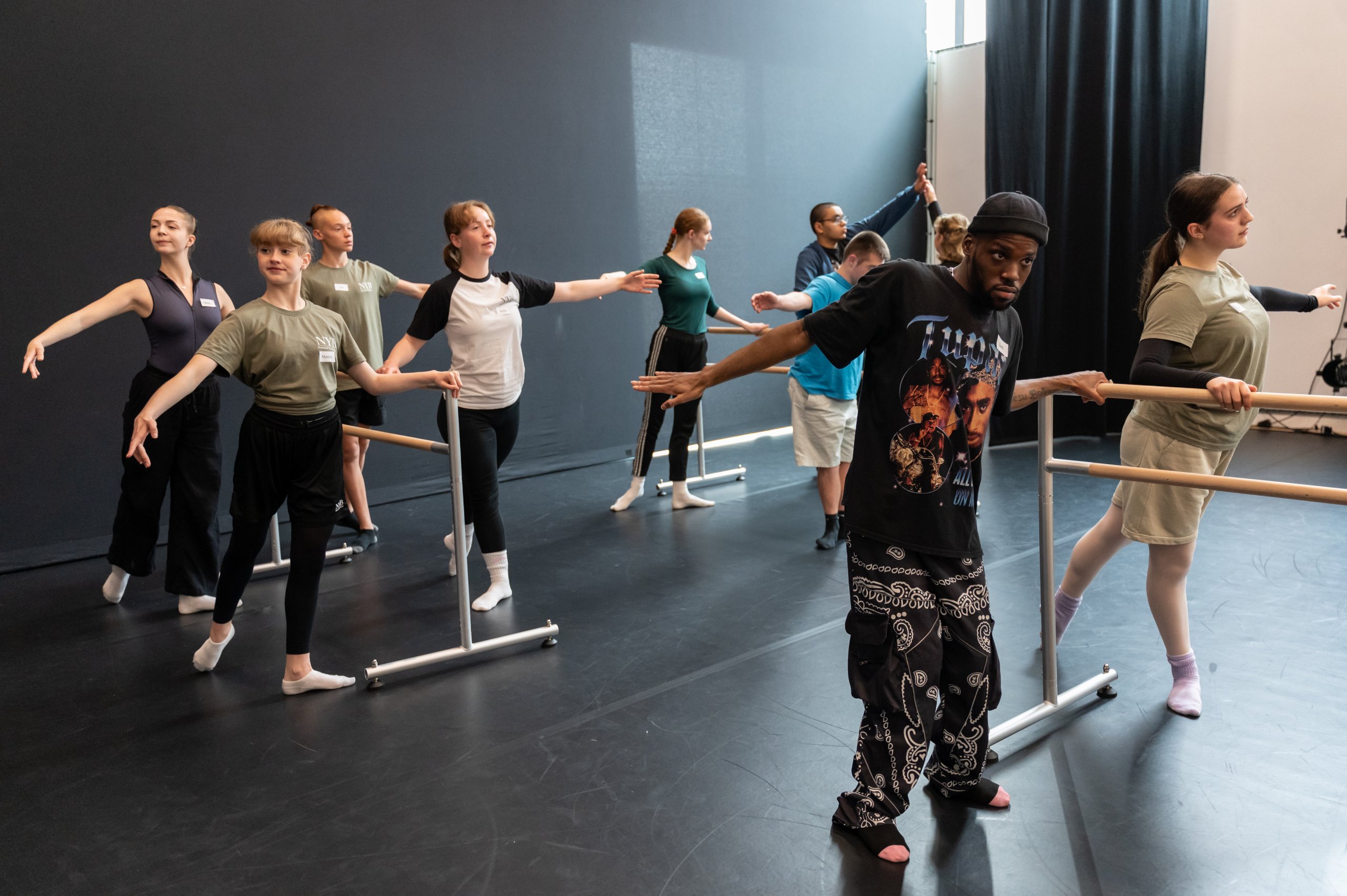 A class of dancers standing and holding onto a ballet barre, some dancers have a leg reaching out behind them. 