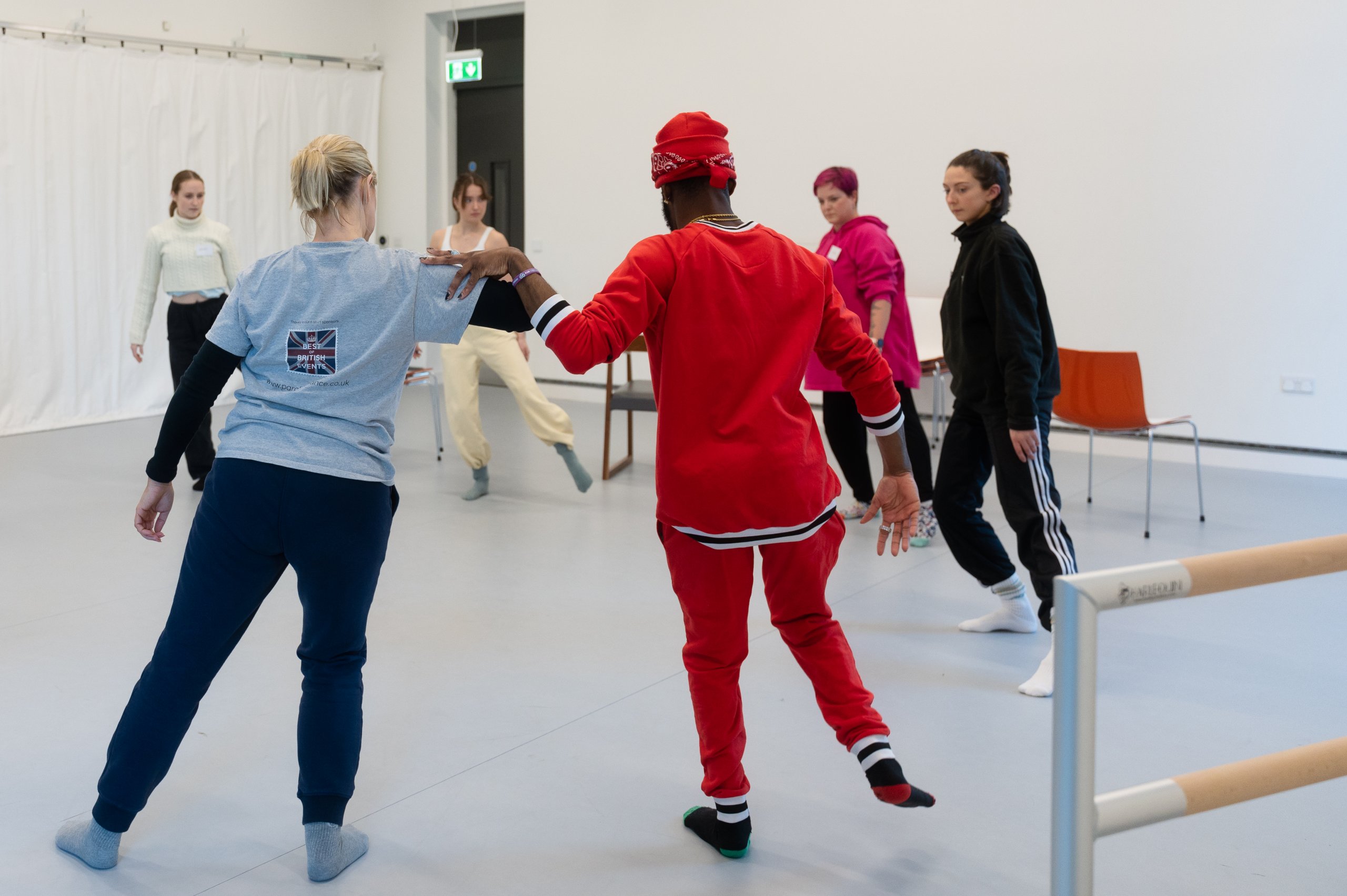Six dancers stand in a circle, transferring weight onto one leg. Two dancers in the centre of the image and making contact with their arms. 