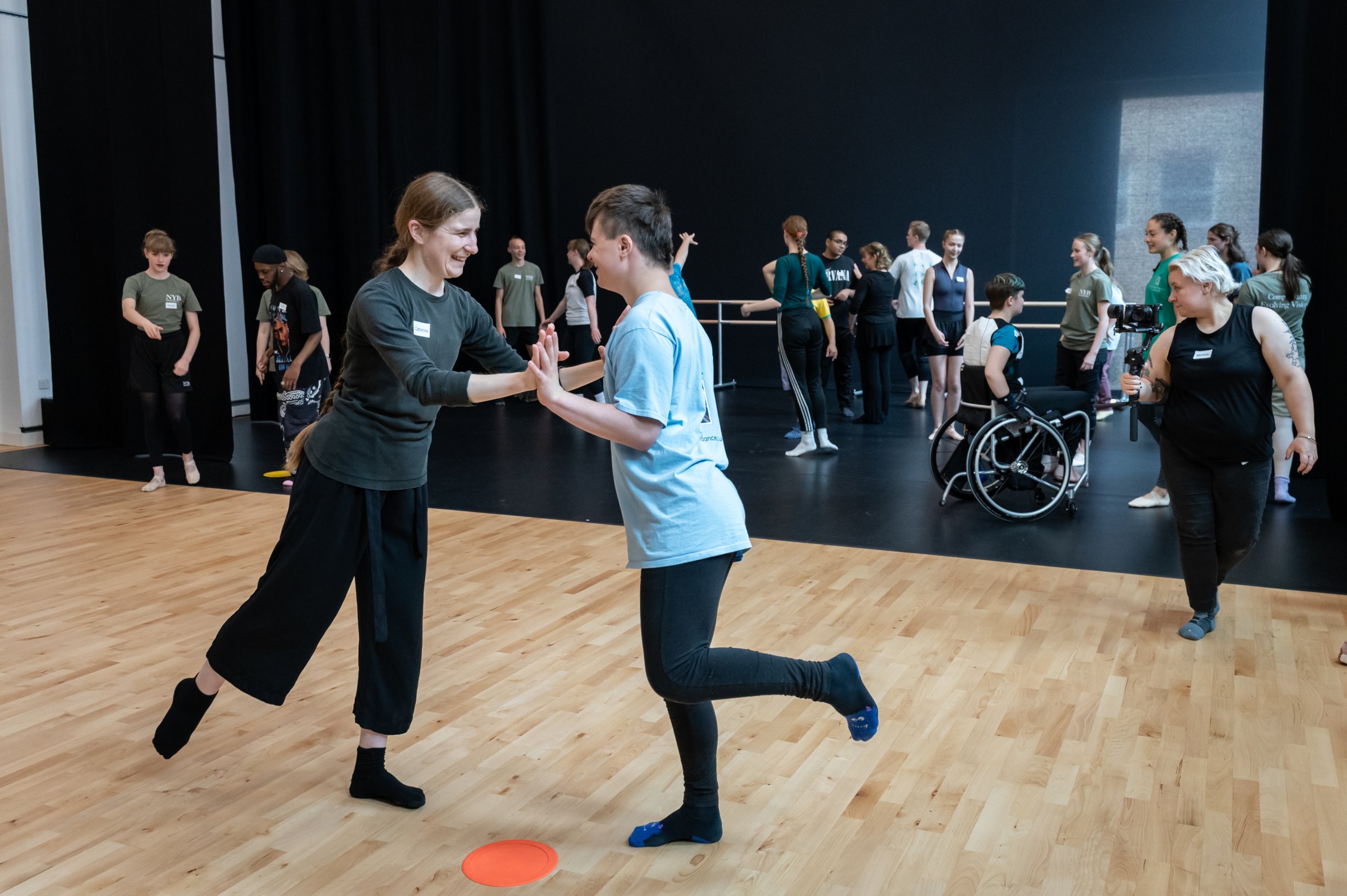 Two dancers stood between an orange circular pad on the floor, on one leg making contant with their hands, facing each other. The rest of the company of dancers are in the background of the image. 