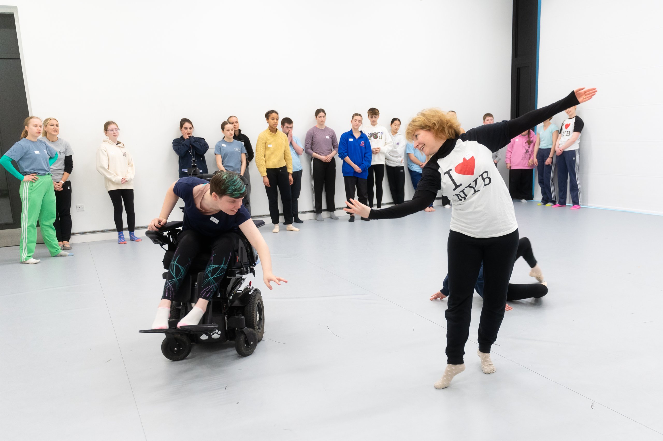 A dancer in a wheelchair reaching towards the floor with one arms, next to a dancer with arms spread wide looking at each other. Several dancers observing in the background. 