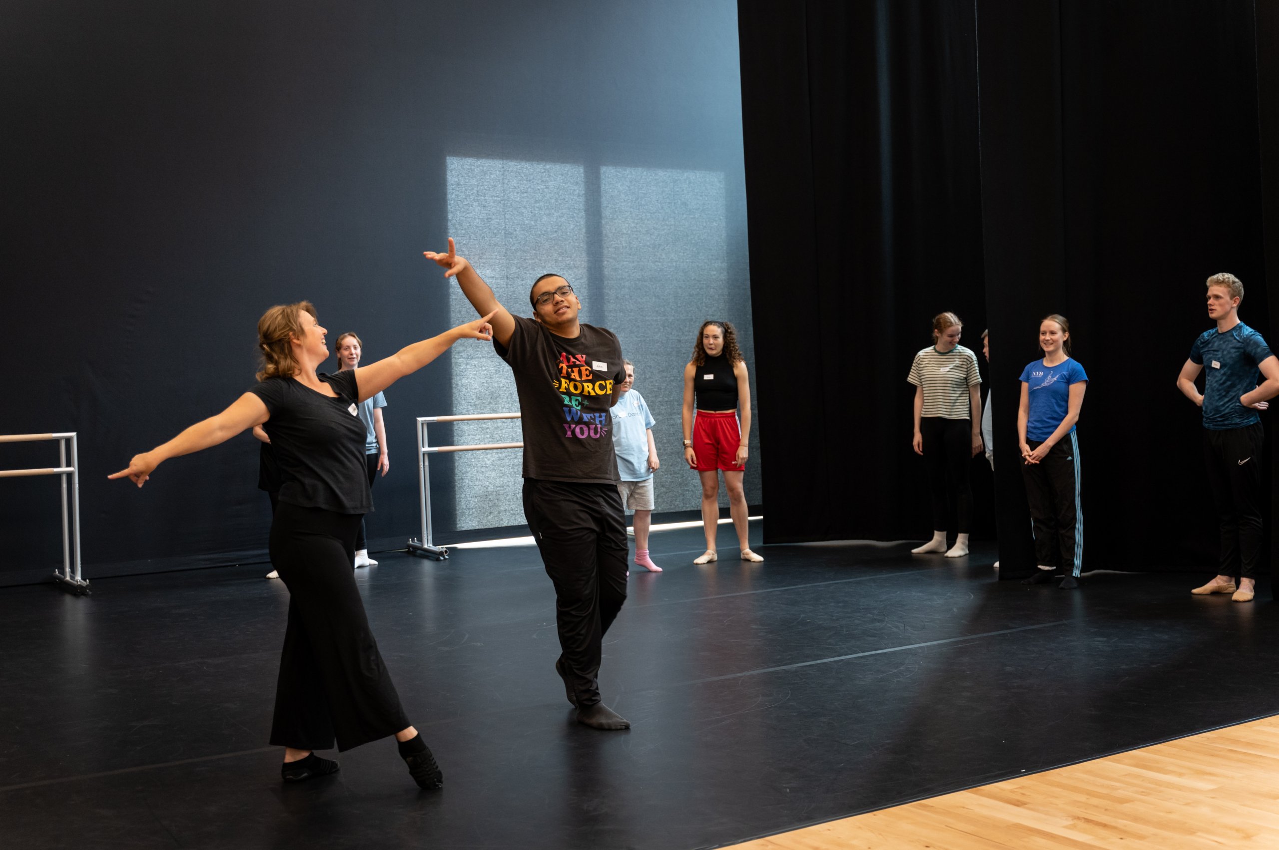 Two dancers reaching with their arms towards each other with several company dancers observing in the background. 