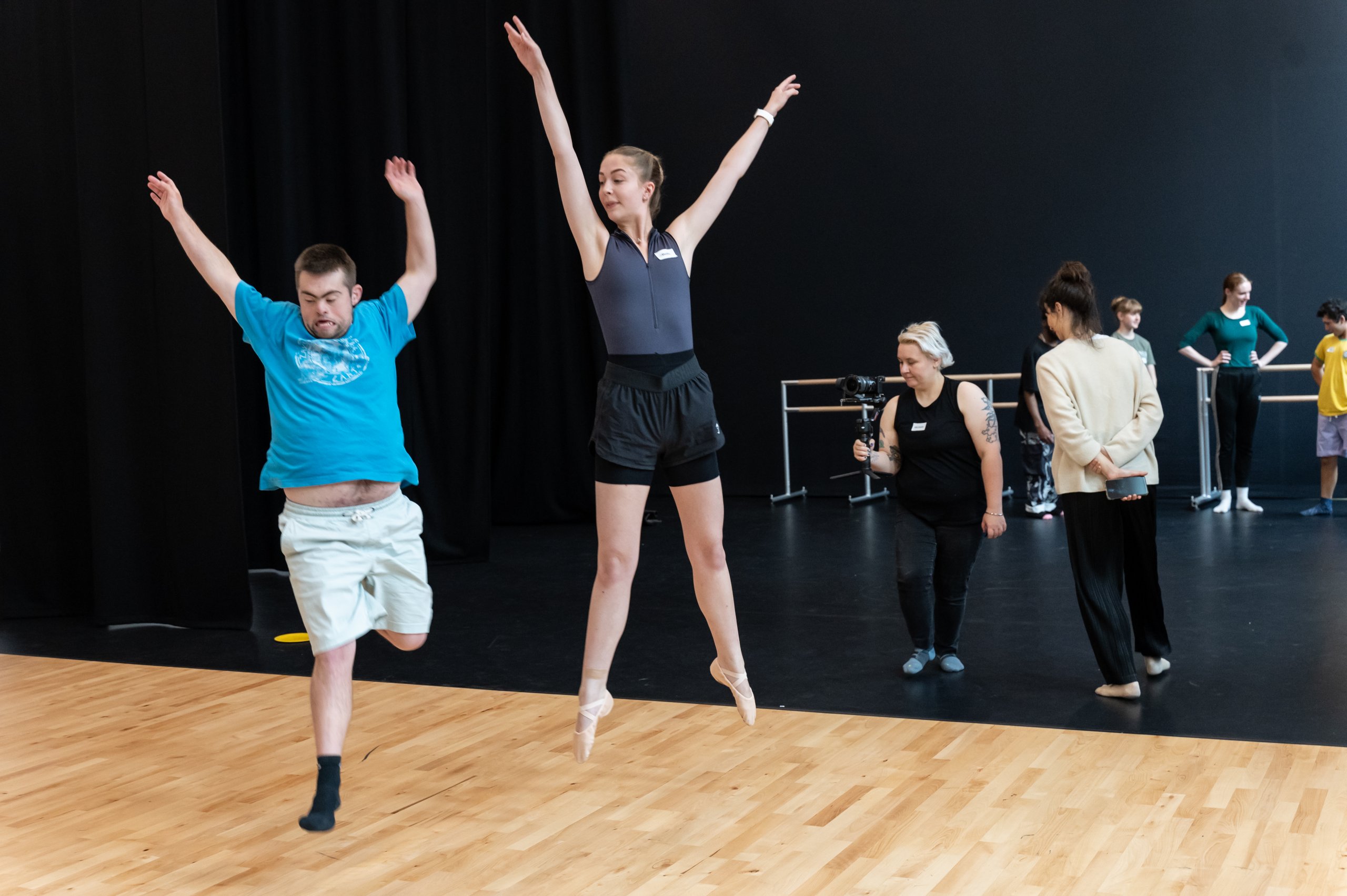 Two dancers elevated off the ground with limbs reaching in different directions. There are some people in the background observing them. 