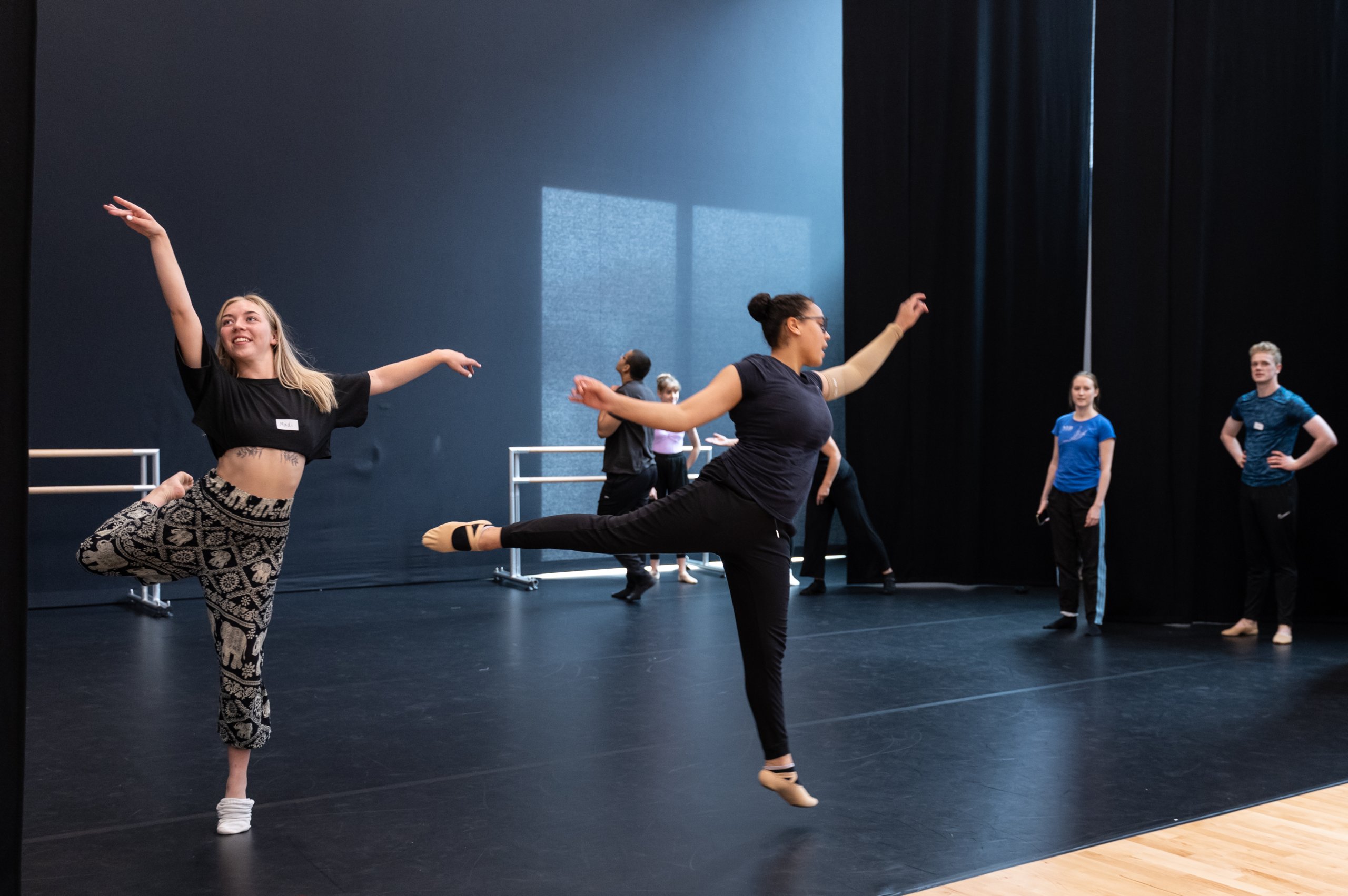Two dancers balance on one leg with the other leg extended to the back and arms extended outwards. Some dancers are observing. 