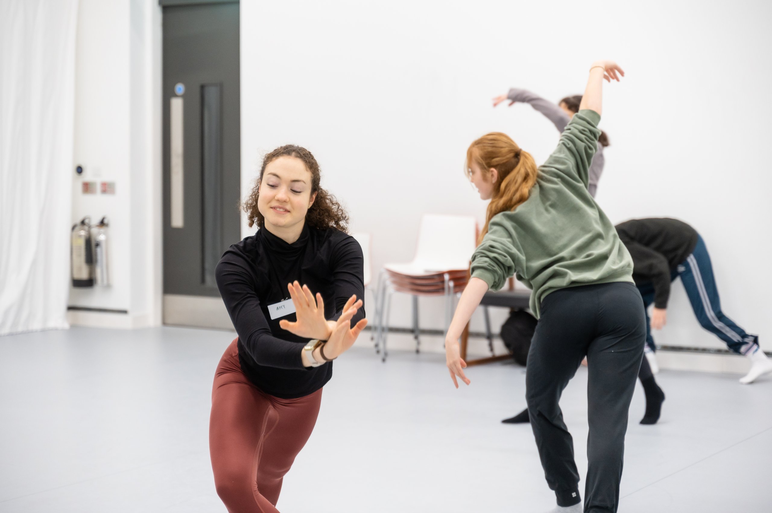 One dancer crossing their wrists whilst lunging whilst another dancer facing the opposite direction reaches arms diagonally in a smaller lunge position. 