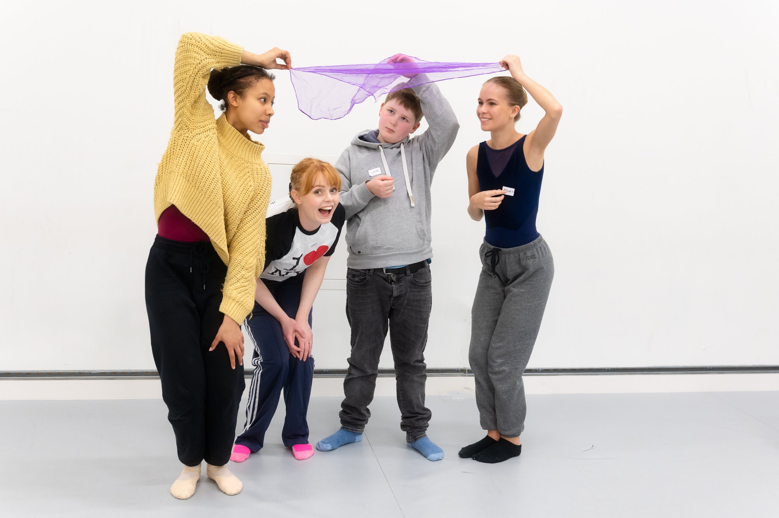 Four dancers beneath a small piece of purple material. Three of these dancers are holding the material and one is crouching beneath. 