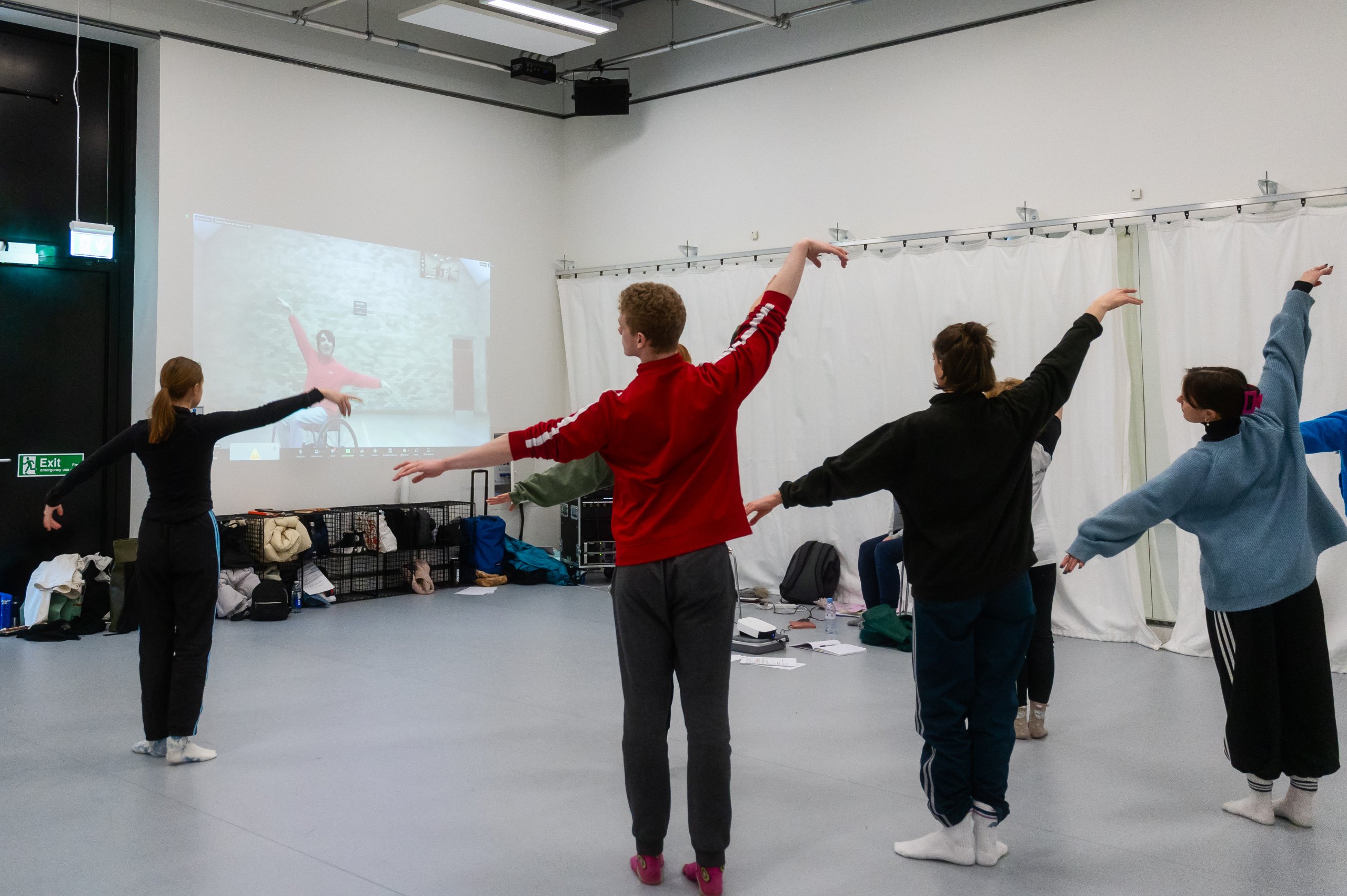 Four dancers with their arms lifted diagonally facing a projection of a dancer in a wheelchair on the wall. 