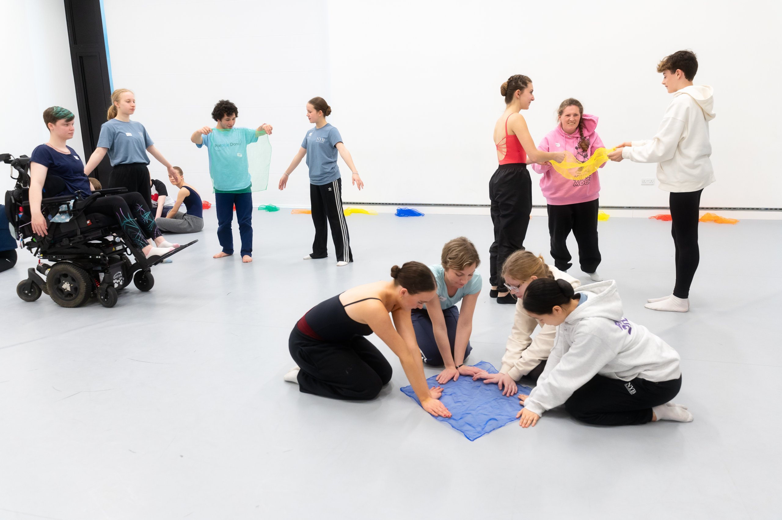 Four dancers in the foreground kneeling with their hands placed on a blue piece of material on the floor. A group of three behind them holding a yellow piece of material and a group of four dancers next to them with arms in a low V postion. One dancer is holding a green piece of material and another dancer is in a wheelchair. 