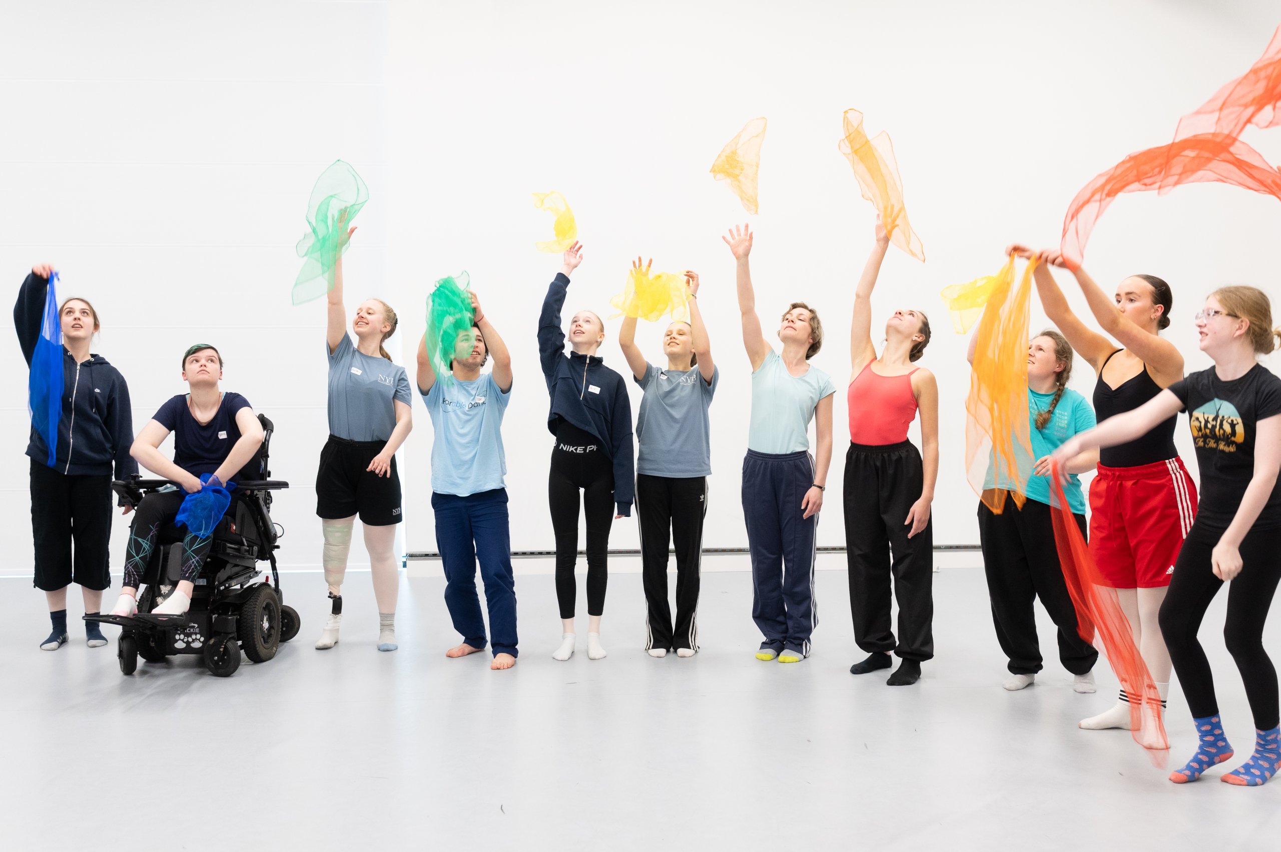 A line of dancers, each holding a colourful piece of material and waving it. 