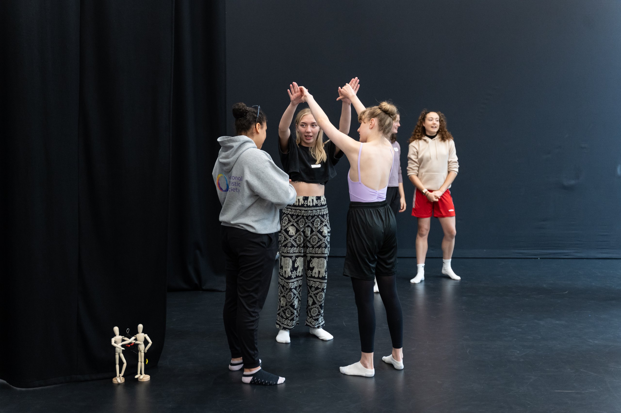 A group of dancers stood in a small circle with two small wooden mannequins on the floor next to them. 