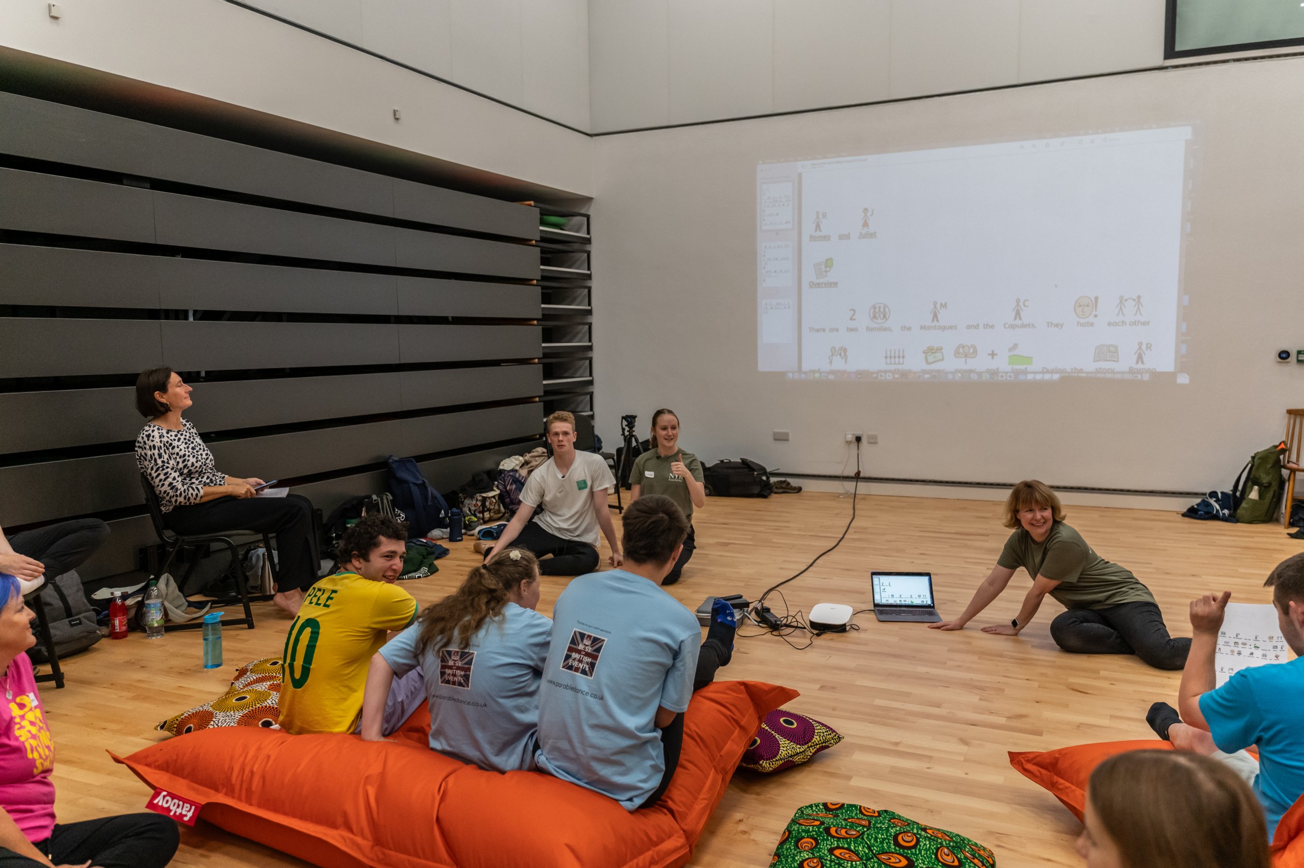 Many people sit on beanbags whilst looking at a projection on the wall. There is a person with a laptop sat facing them. 