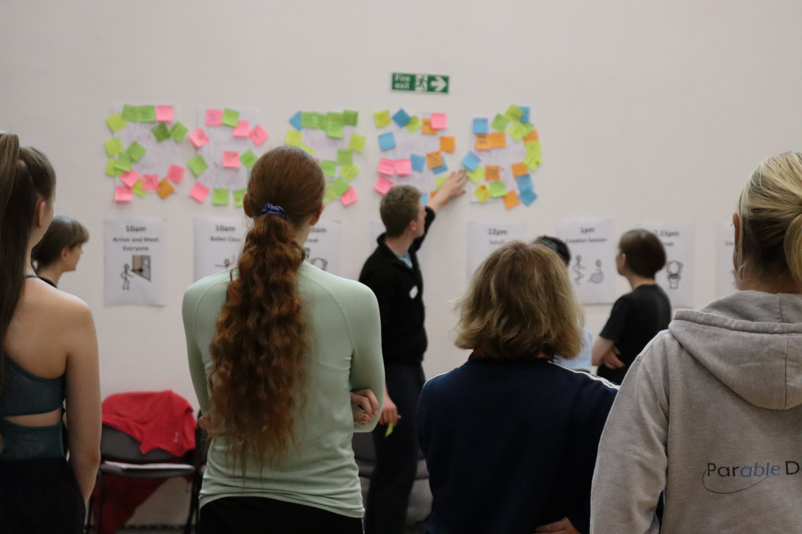 A person pointing at colourful post stick notes on a wall whilst several people are observing this. 