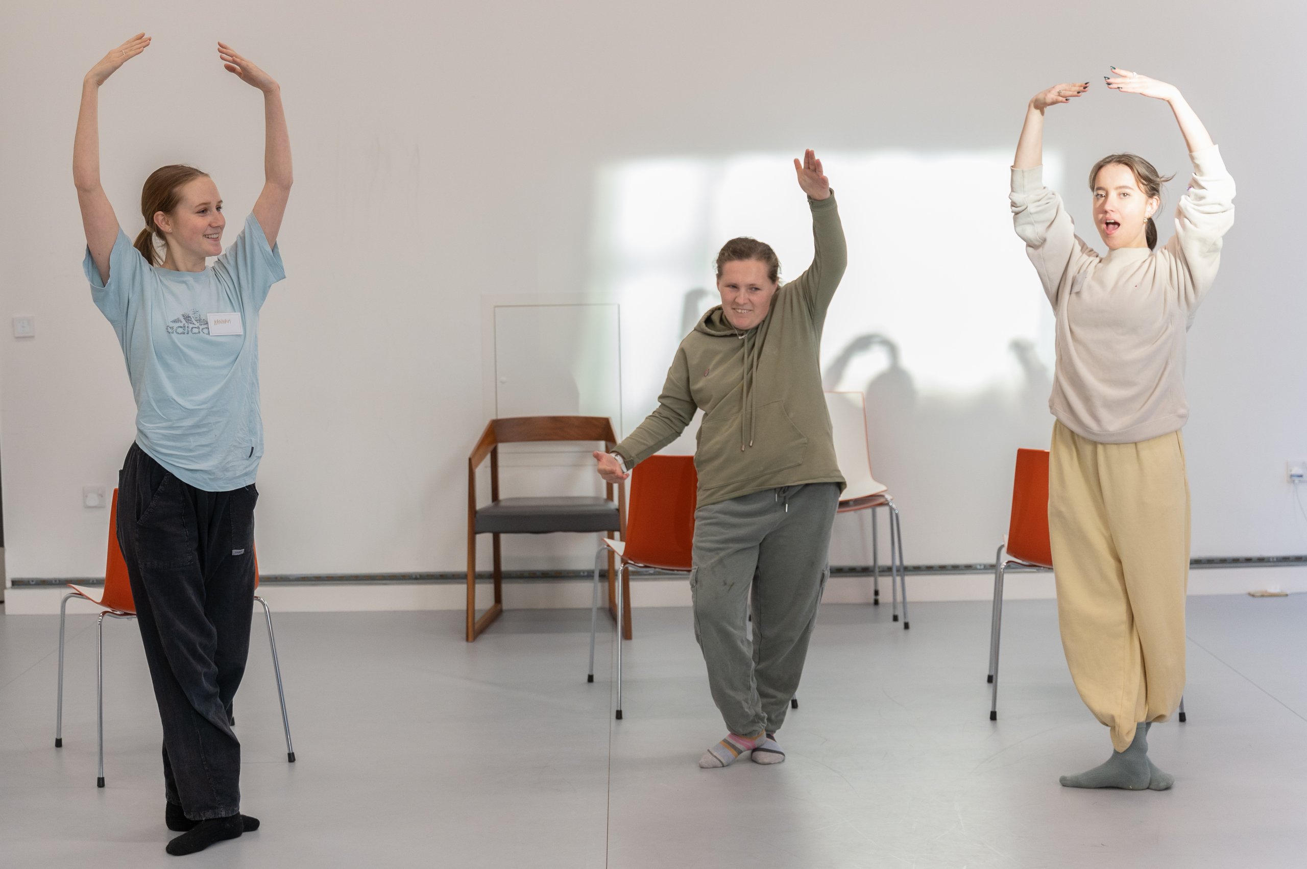 Three dancers standing with either one or two arms raised above their heads. There are three red chairs behind them. 