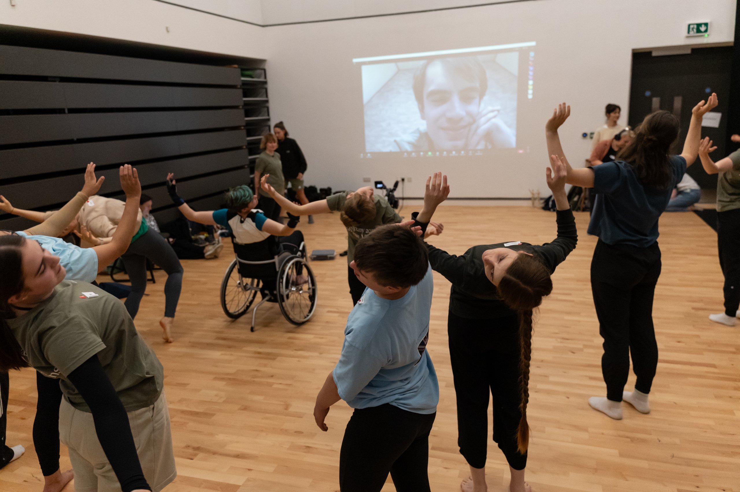 Many dancers with arms raised in a leaning backwards motion with a person projected onto the wall. 