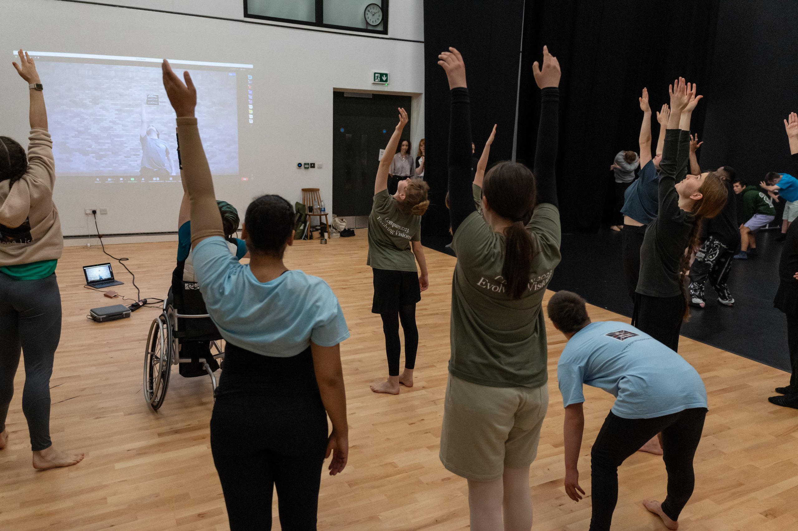 Several dancers reaching upwards with one or two arms whilst one person reaches down to the floor. There is a projection on the wall. 