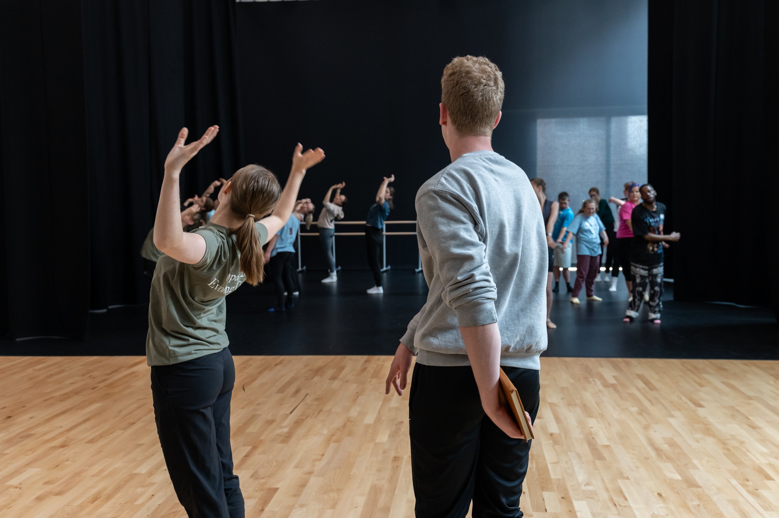 One dancer with arms bent and raised at the side leaning backwards next to another dancer twisting their spine and arms. There are many dancers observing this. 