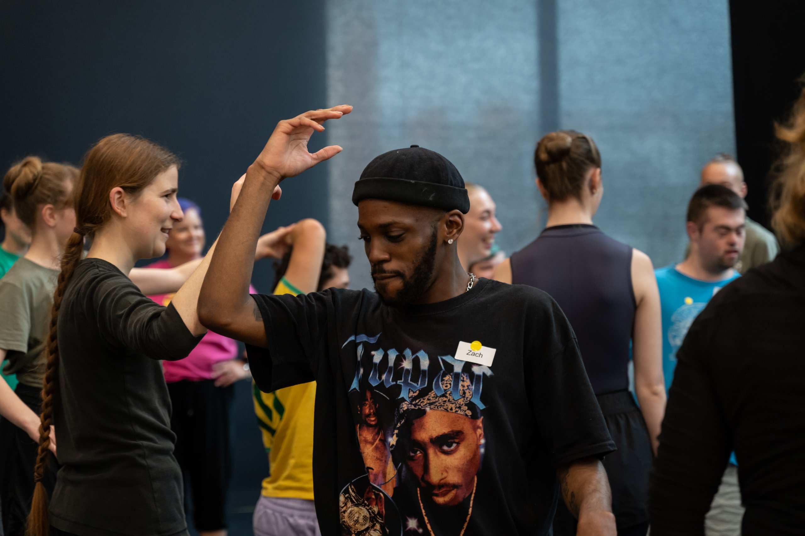 A dancer in a black t-shirt and hat lifting one arm so the elbow and shoulder are in line. There are many dancers in the background in motion. 