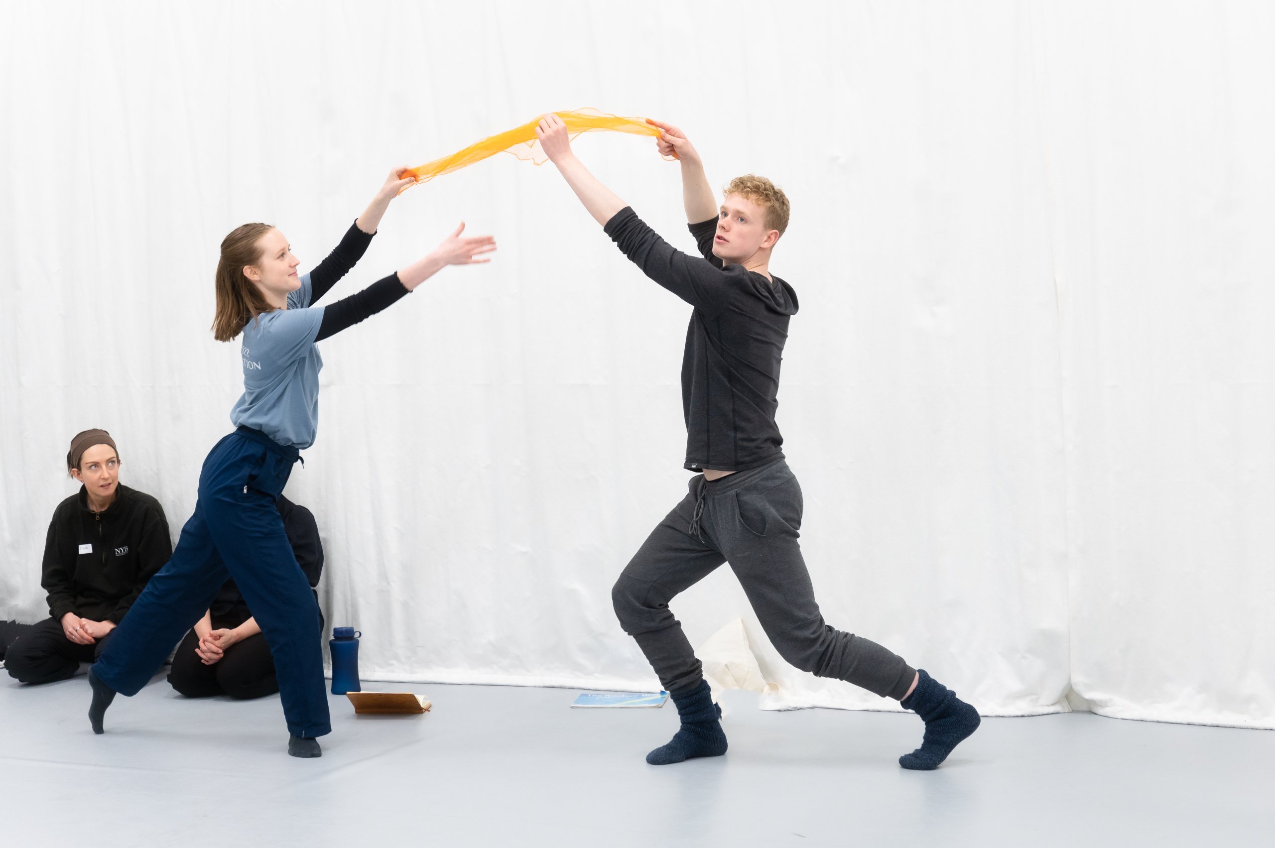 Two dancers lunging towards each other whilst both holding a yellow piece of material whilst two people are sat on the floor observing them. 