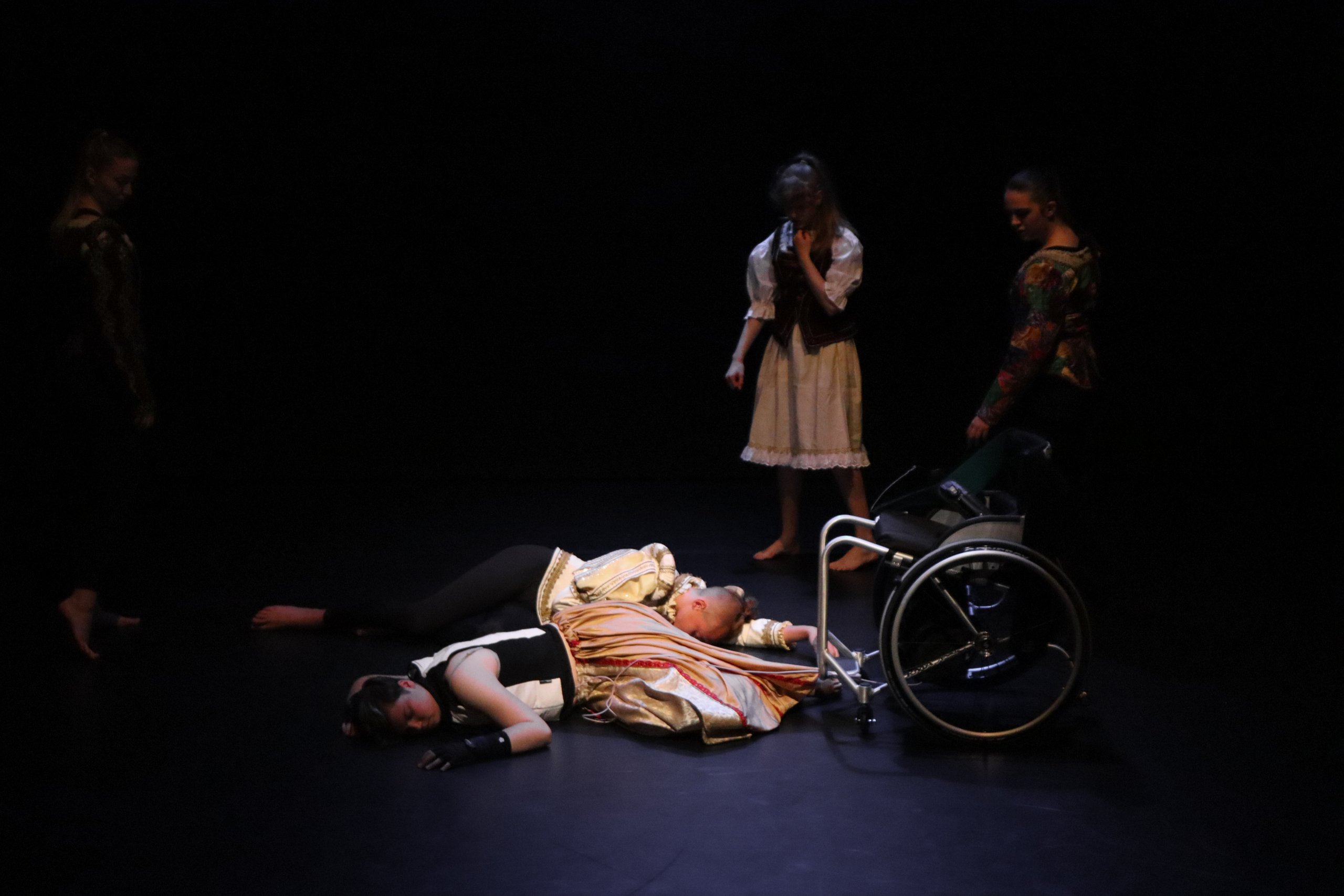 Romeo and Juliet laid on the floor with one dancer looking down over them, lit slightly by the stage lighting. 