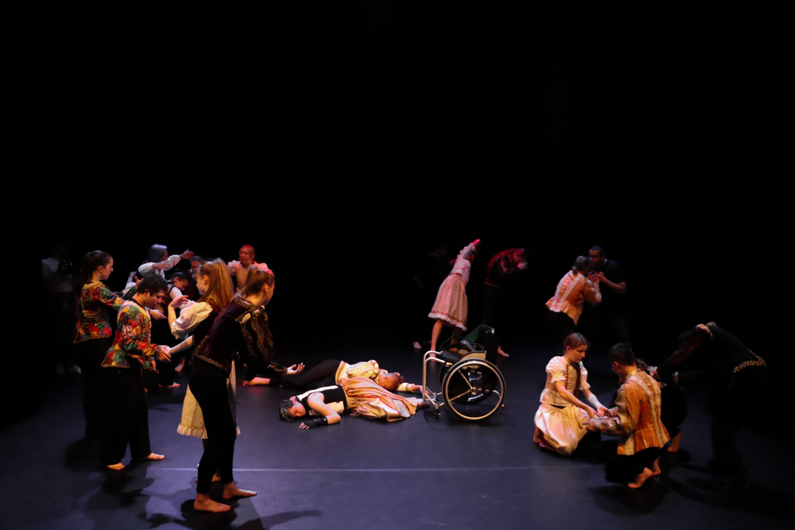 Dancers on stage in costume surrounding Romeo and Juliet who are laid on the floor with a wheelchair placed next to them. 