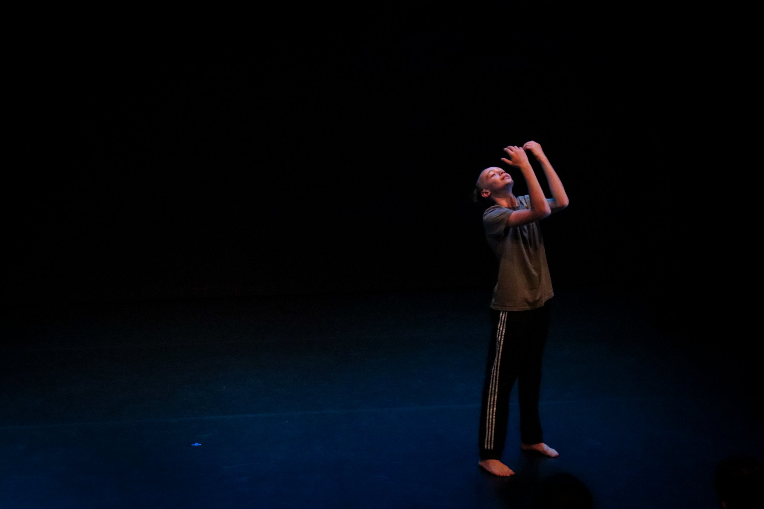 One dancer in a dark space in parallel, looking upwards with elbows bent and hands in front of their face. 