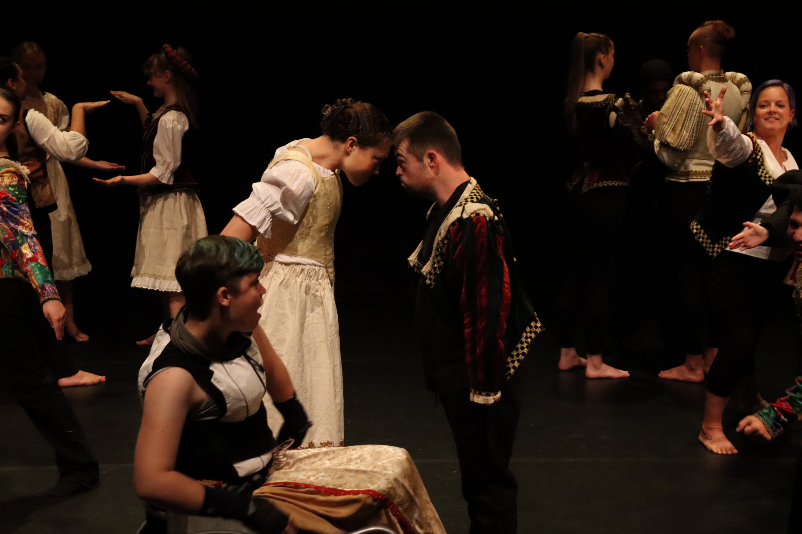 A dancer in a wheelchair is in front of two dancers connected at their foreheads with several other dancers behind them in motion. 