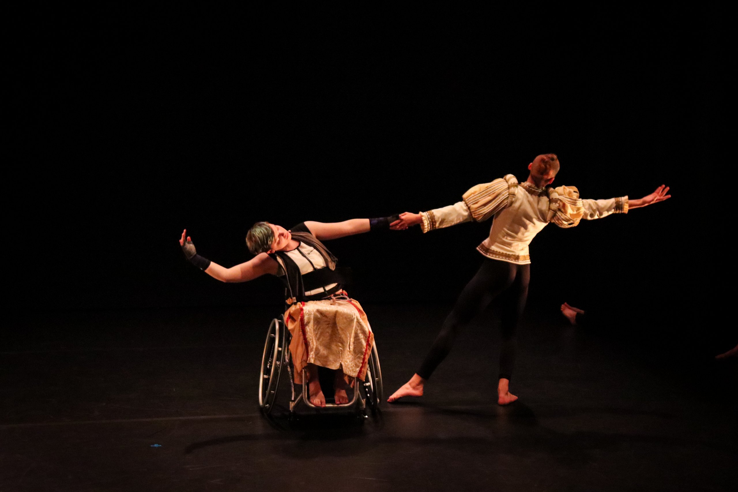A dancer in a wheelchair holding hands and leaning away from a dancer standing. 