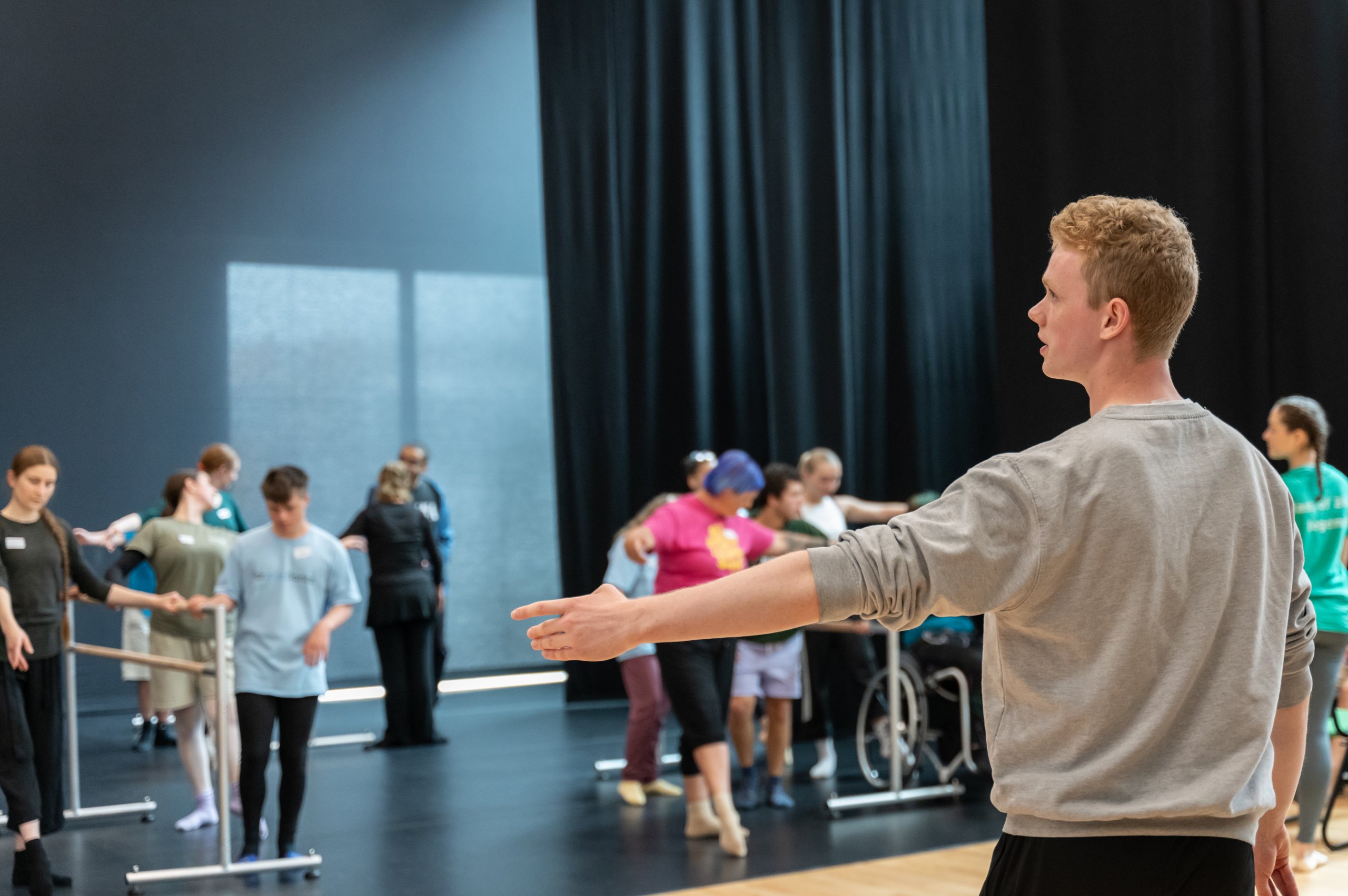 Ballet teacher is demonstrating with their arm extended out to the side. In the background the participants are observing this demonstration. 