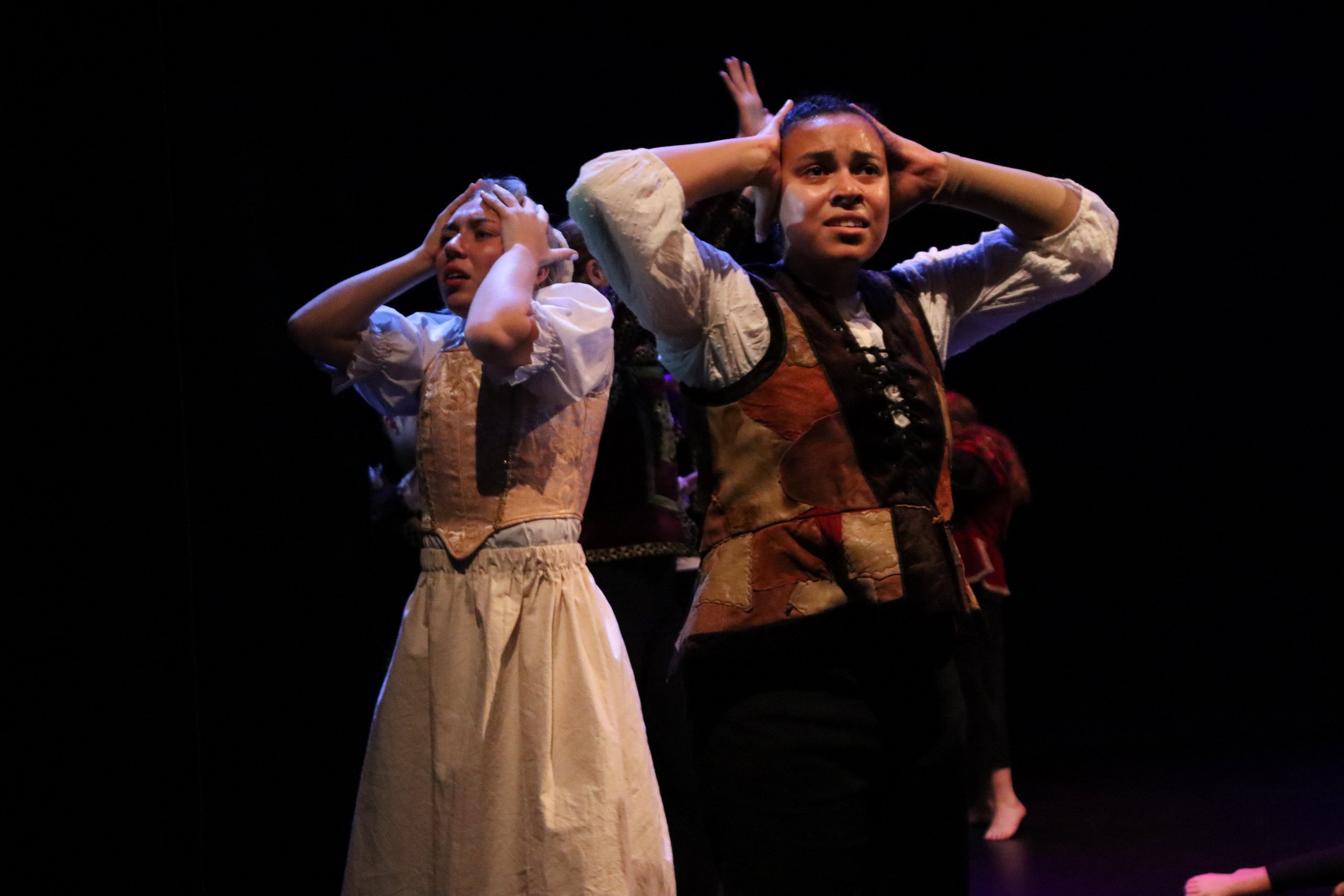 Two dancers in costume with hands on the side of their heads with elbows pointed out to the side with expressive faces. 