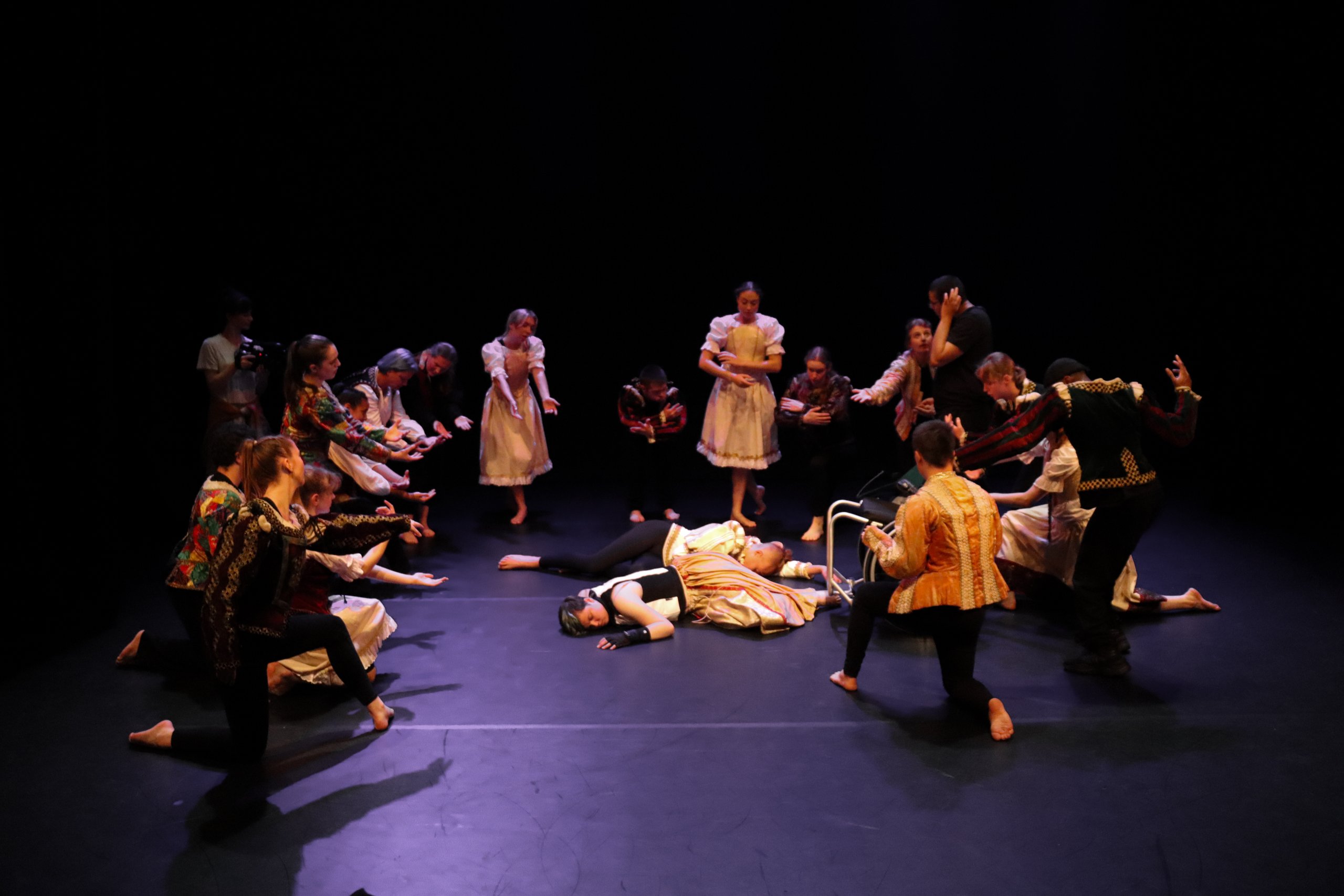 Dancers on stage in costume surrounding Romeo and Juliet who are laid on the floor with a wheelchair placed next to them. 