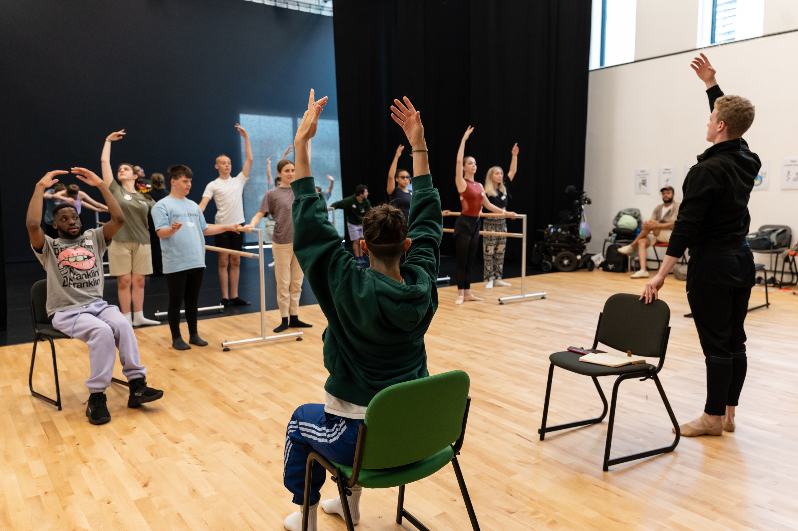 Dancers in varying positions. Some dancers are setaed on chairs and others standing and holding onto a ballet barre with arms extended with either one or two arms in fifth position. 