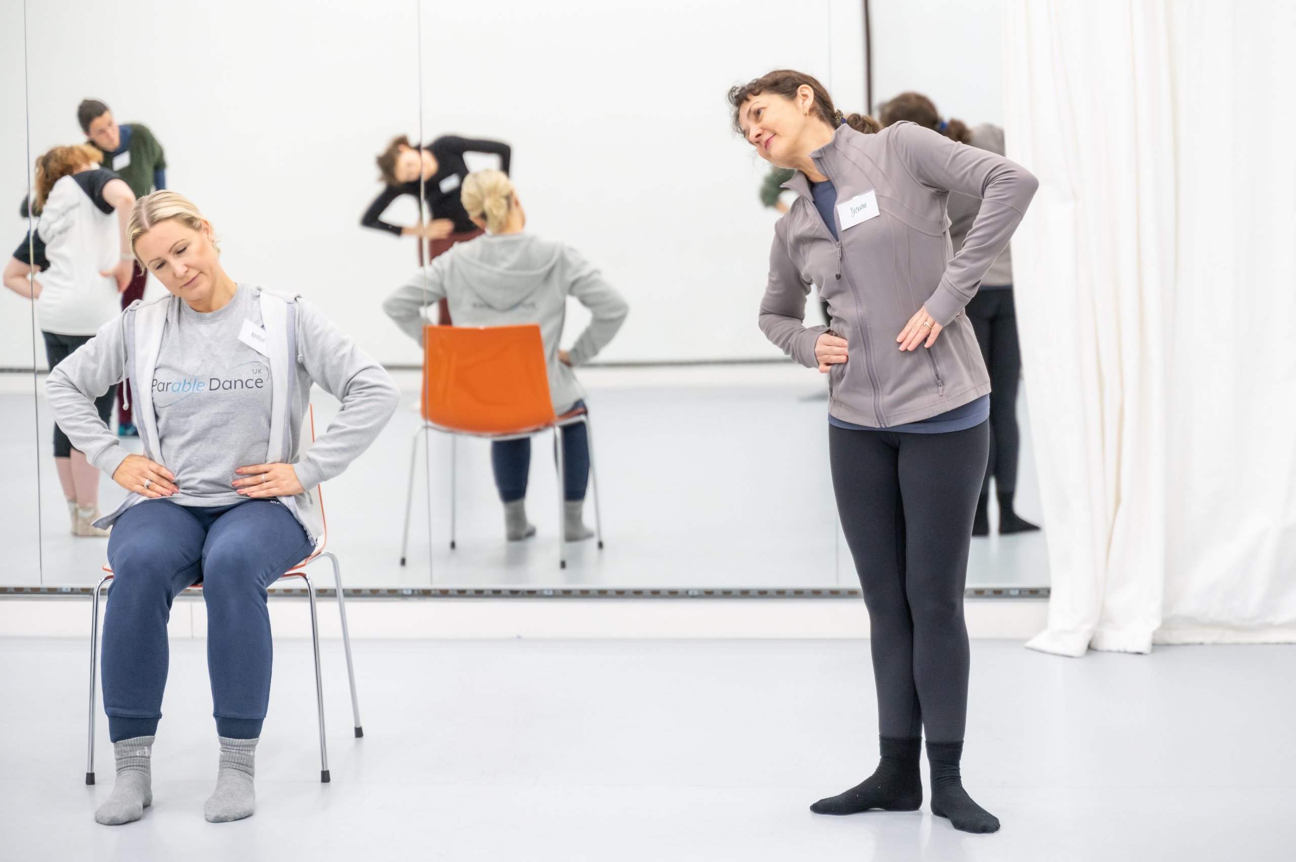 A dancer seated on a chair with arms placed on their hips and head tilted to one side next to a dancer standing in first position with hands on their hips with their head tilted. 